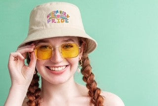 Person wearing a beige bucket hat with 'Museum Nerd Pride' colorful text and yellow sunglasses on a light green background