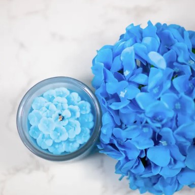 An image of a blue flower-shaped candle in a clear container next to a bouquet of blue flowers on a white background