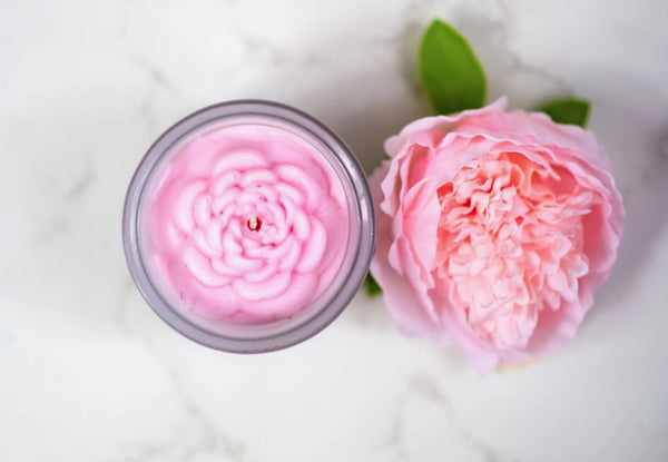 Pink flower shaped candle in a jar next to a pink flower on a white background