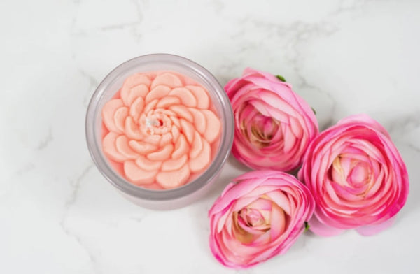 An image of a pink flower-shaped candle in a clear container next to a trio of pink flowers on a white marble surface