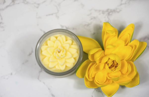 Yellow flower-shaped candle next to a small glass container with a floral design on a marble surface